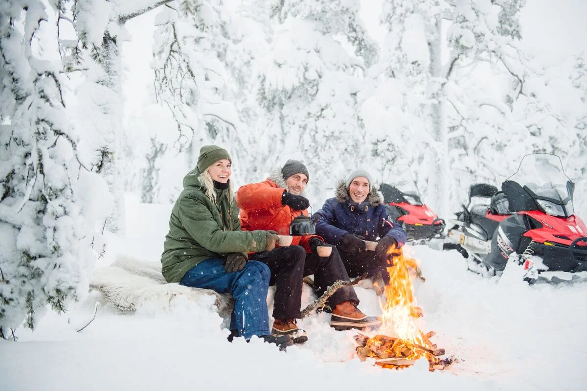 Three colleagues warming up around a campfire in deep snow, snowmobiles parked behind them
