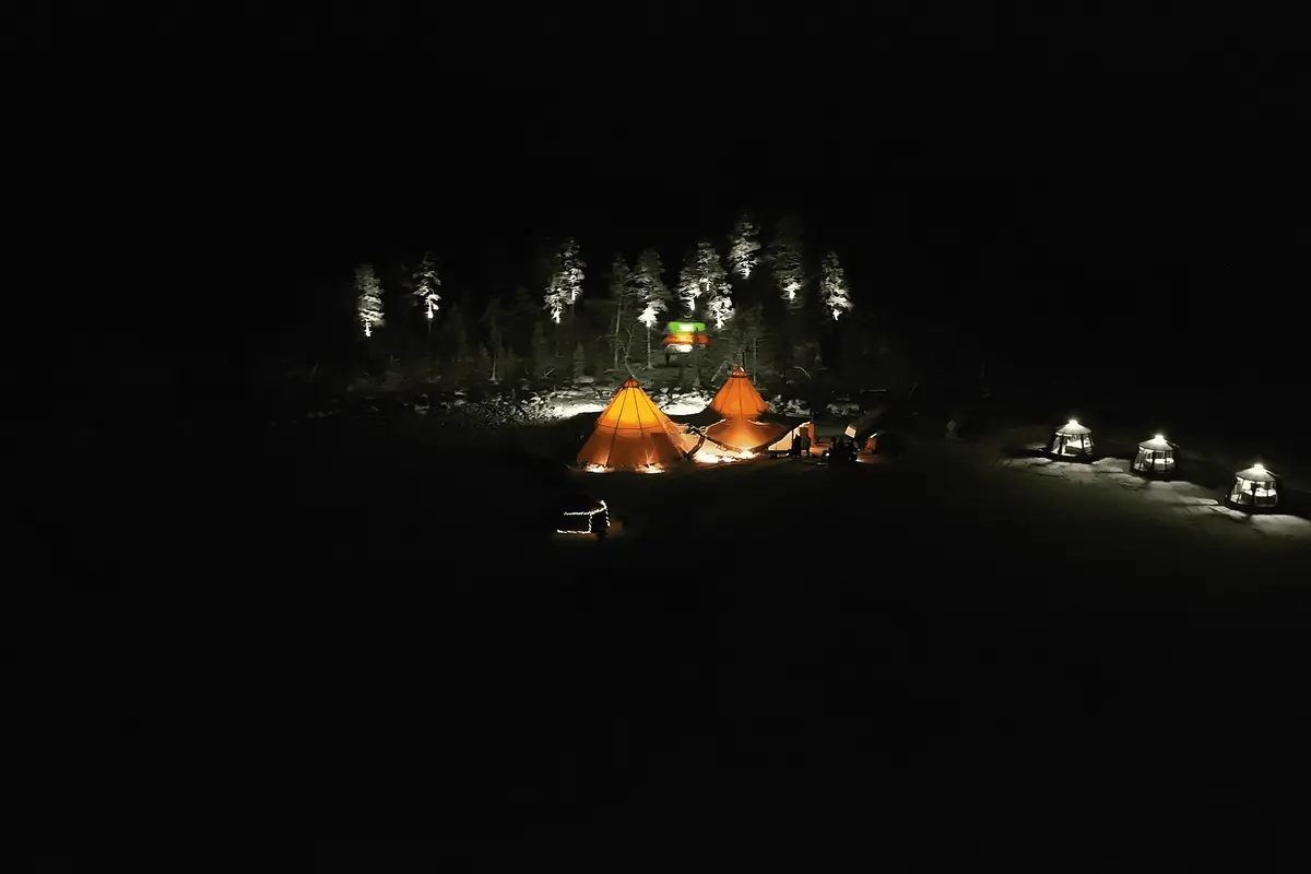 Two lit lavvu tents and a row of glass igloos in a dark Lapland forest clearing, seen from the air at night