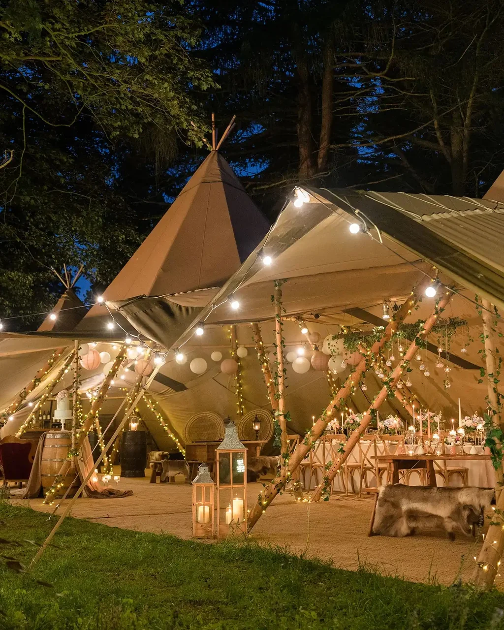 Twin Tentipi Stratus 72 Nordic event tipis joined and lit with festoon string lights at twilight, candle lanterns and reindeer hides at the entrance, wooden banquet tables glowing inside — a Lapland wedding venue ready for guests